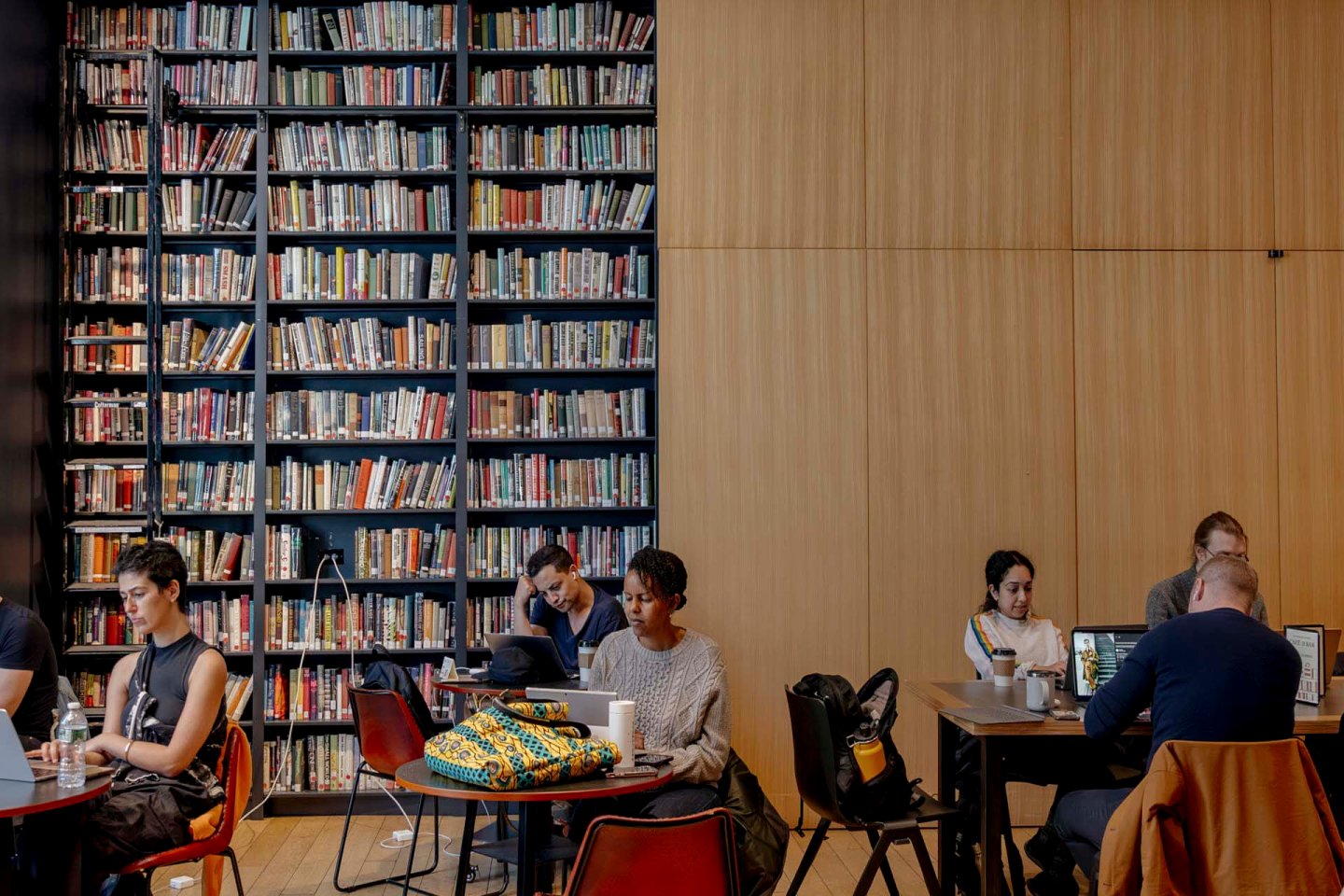 People studying and working on laptops at tables in a library. The room features a large wall filled with bookshelves and another wall with wooden paneling. Some individuals are reading while others are focused on their screens.