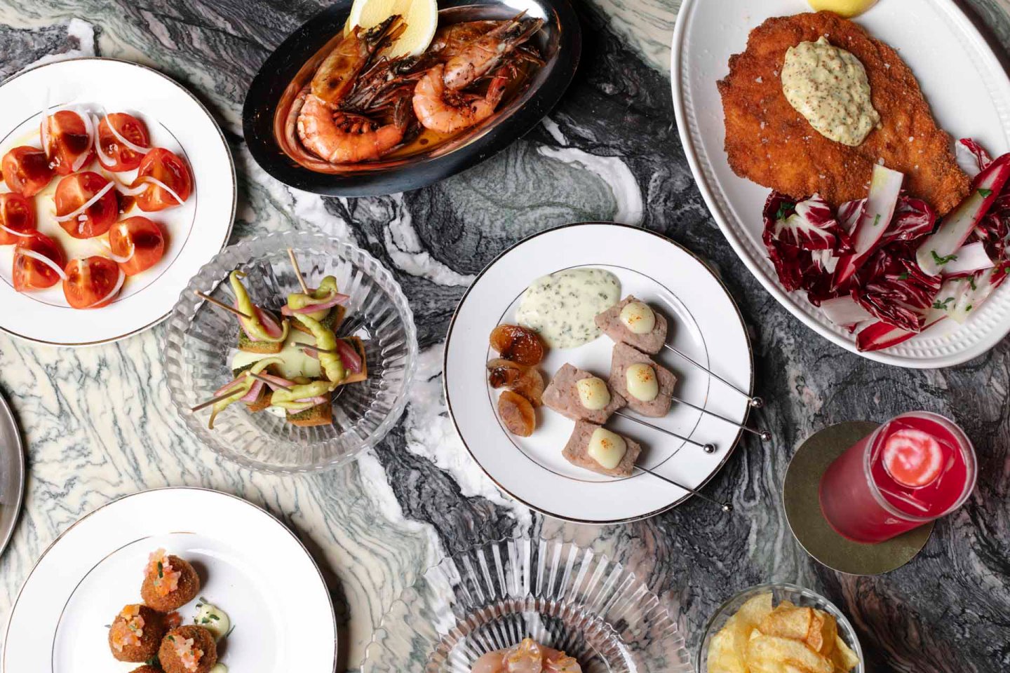 A variety of dishes on a marble table, including shrimp with lemon, skewered scallops with sauce, sliced tomatoes and mozzarella, breaded meat with salad, fried bites, artichoke salad, and a pink drink.