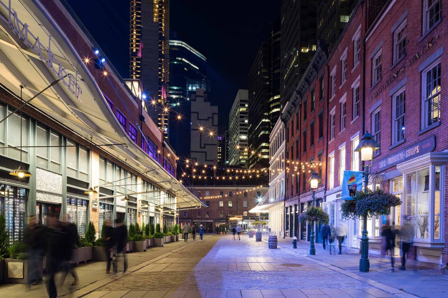 A lively city street at night features string lights overhead, historic brick buildings on the right, modern buildings on the left, and blurred pedestrians walking along the illuminated sidewalk.