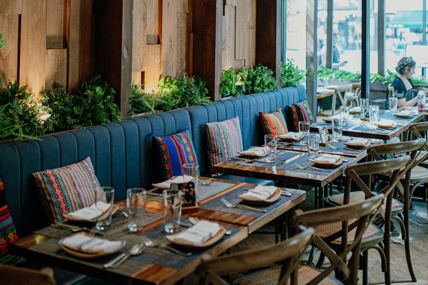 A cozy restaurant interior with wooden tables set for dining, blue cushioned bench seating, colorful decorative pillows, and green plants against a wooden wall, with large windows letting in natural light.