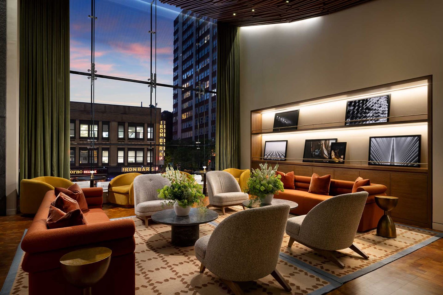 Modern lounge with orange sofas, gray chairs, and floral arrangements on tables. Large windows show city buildings at sunset. Photos and lights decorate the back wall above the seating area.