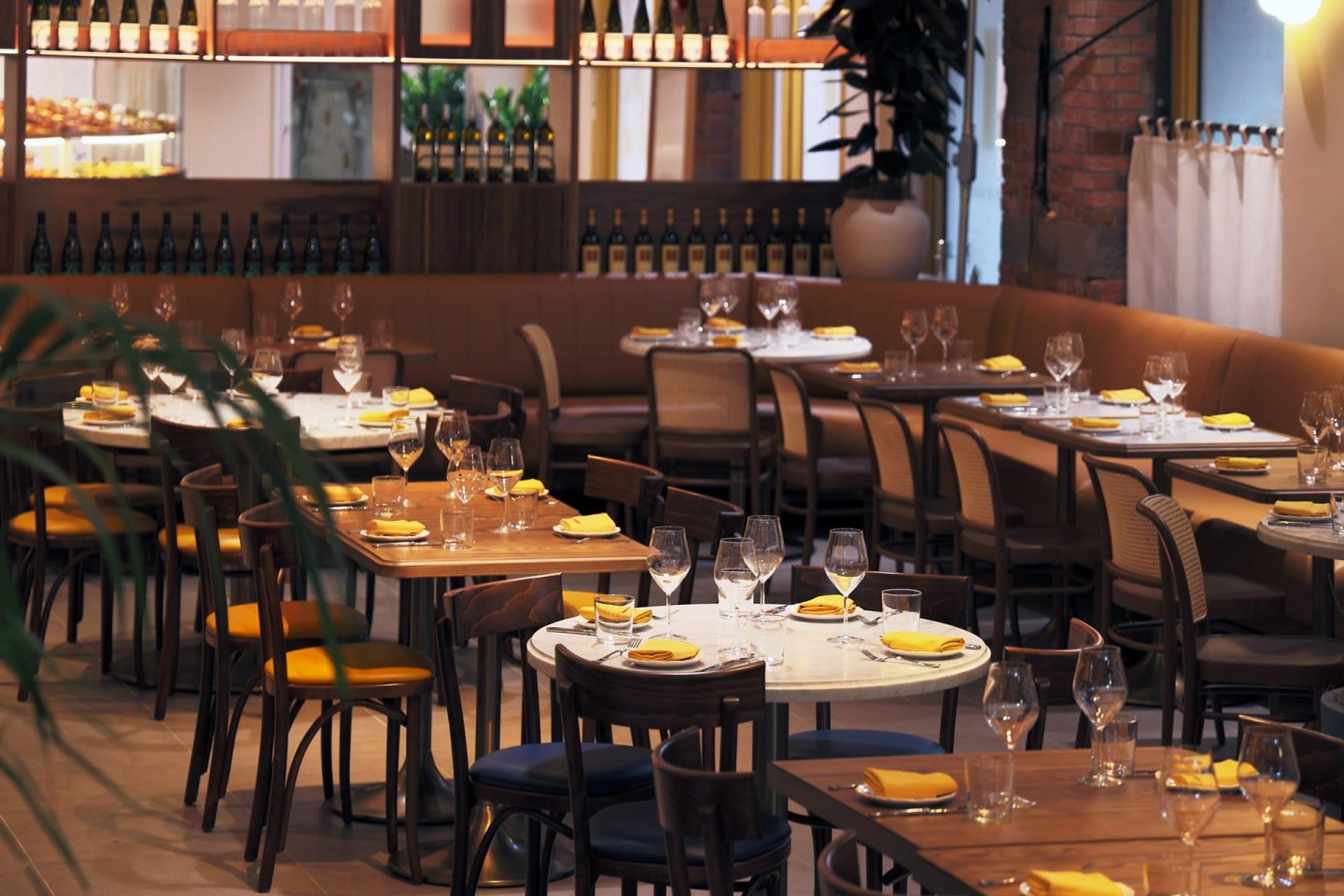 A modern restaurant dining area with neatly set tables, yellow napkins, empty wine glasses, wooden chairs, and a soft, ambient lighting. Wine bottles are displayed on shelves and plants add greenery to the space.