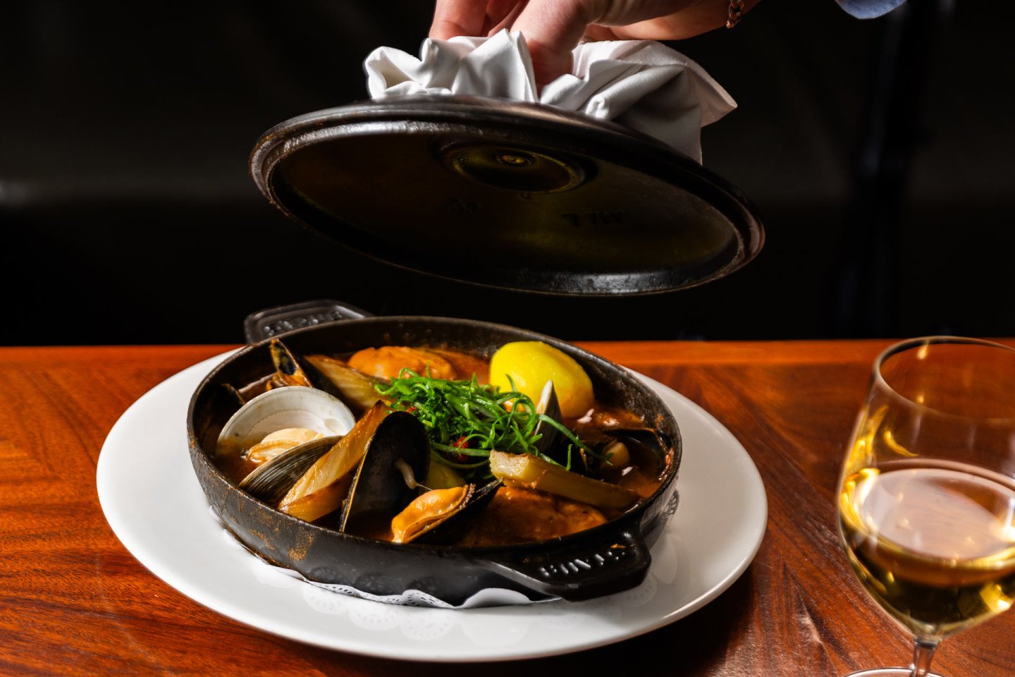 A hand lifts the lid off a cast iron skillet filled with seafood stew, including mussels, clams, and shrimp, garnished with greens and a lemon wedge, served on a white plate next to a glass of white wine.