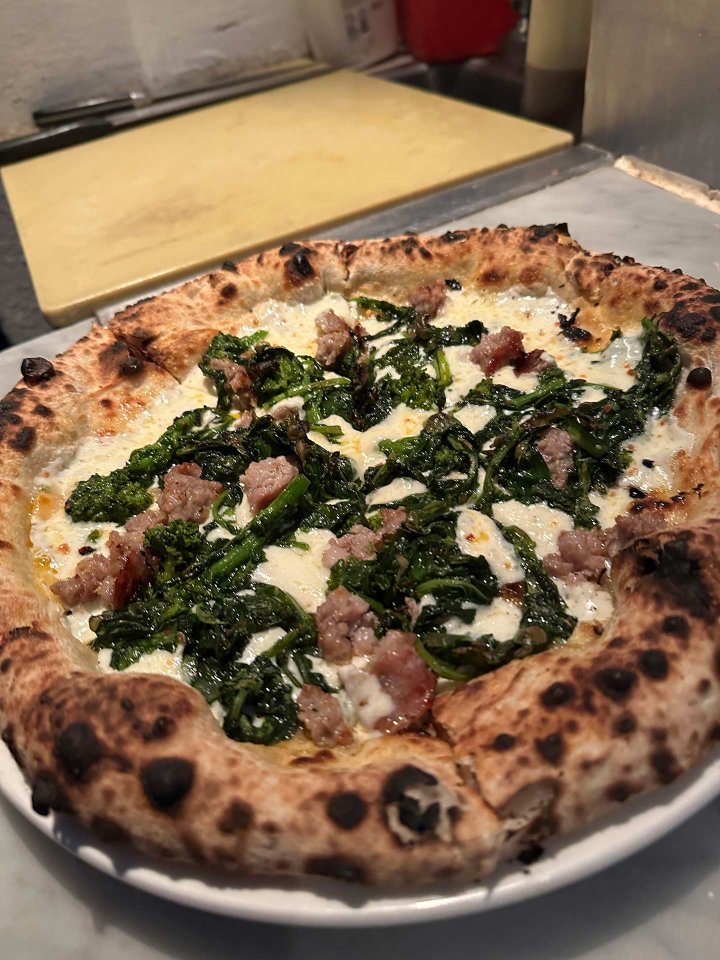 A close-up of a wood-fired pizza topped with broccoli rabe, crumbled sausage, and melted cheese, served on a white plate on a marble counter.