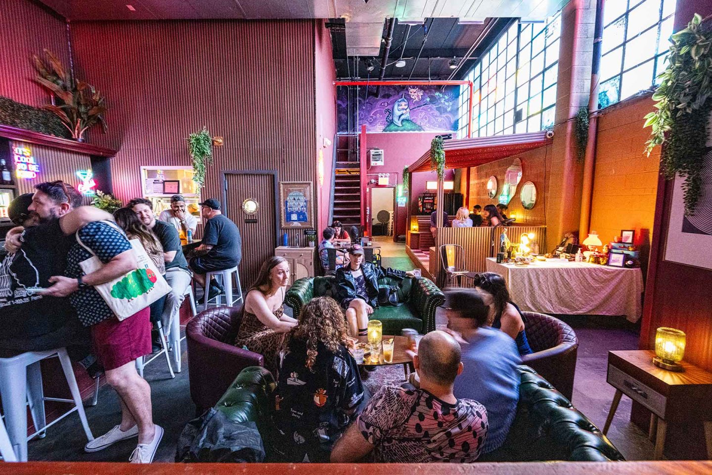 A lively bar with people socializing on couches and bar stools. Colorful lights, large windows, plants, and eclectic decor create a vibrant, cozy atmosphere. A mural is visible above a red staircase in the background.