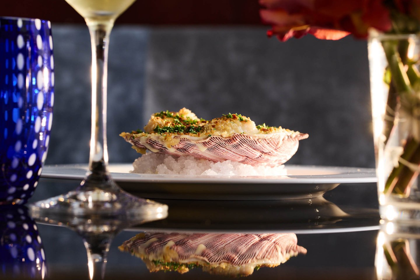 A baked seafood dish garnished with herbs sits on a bed of salt on a white plate, surrounded by a blue glass, wine glass, and a vase of flowers.