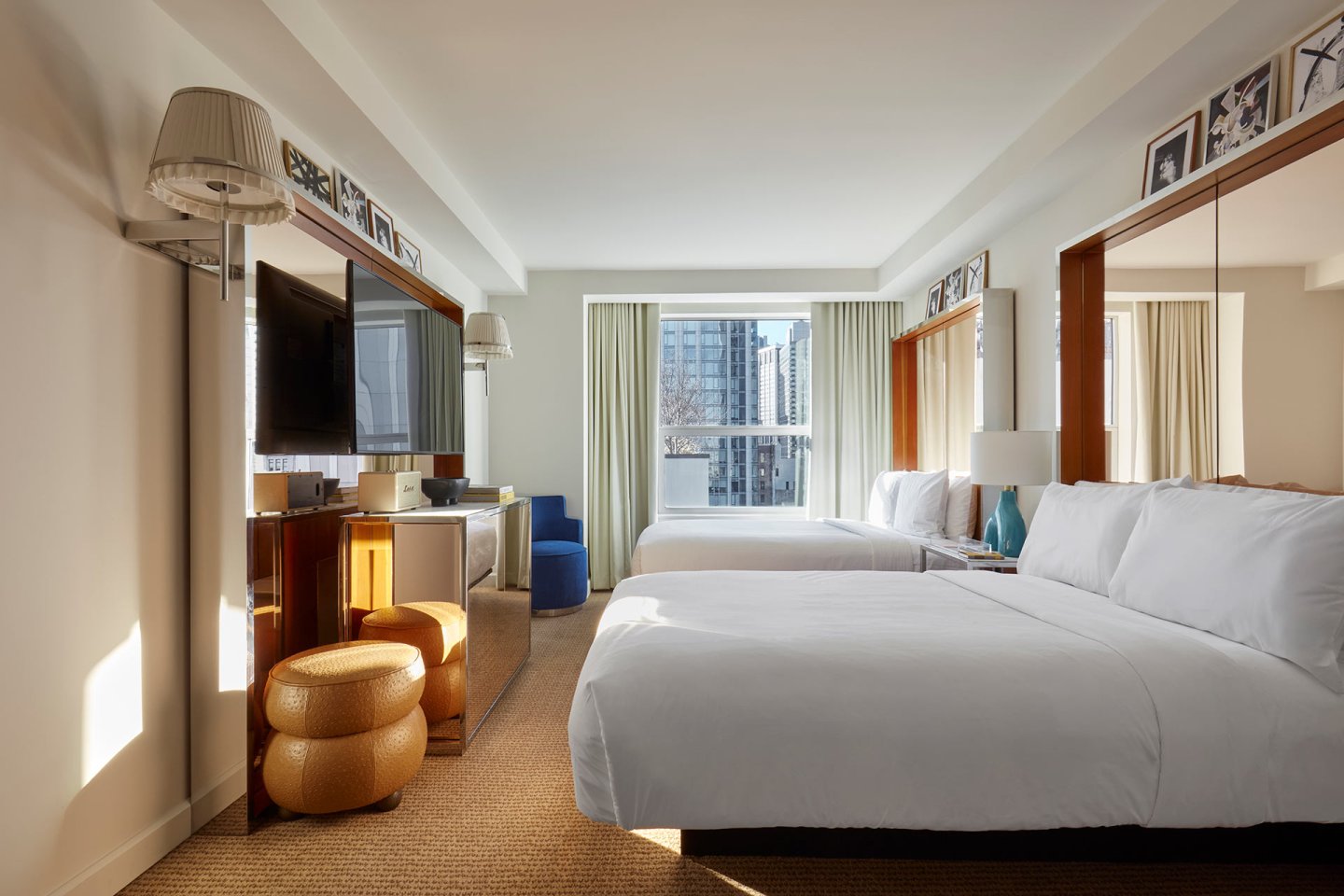 Modern hotel room with two large beds, white bedding, a wall-mounted TV, minibar, windows with city views, and neutral decor. Natural light fills the space, and framed photos are displayed above the beds.