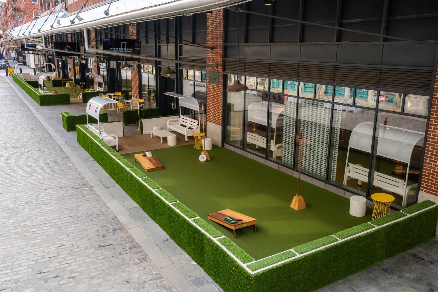 A row of outdoor seating areas with artificial grass, benches, swings, and tables is set up along a brick building on a cobblestone street, separated by green barriers.