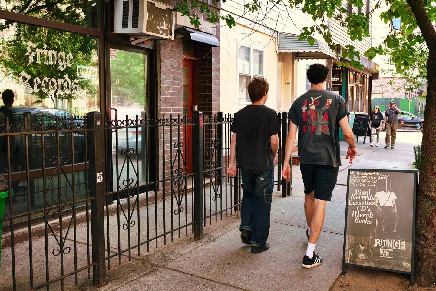Two people walk on a sidewalk past a store called Fringe Records. The storefront has a sign and a display board outside advertising records, cassettes, CDs, merch, and books. Other pedestrians are in the background.