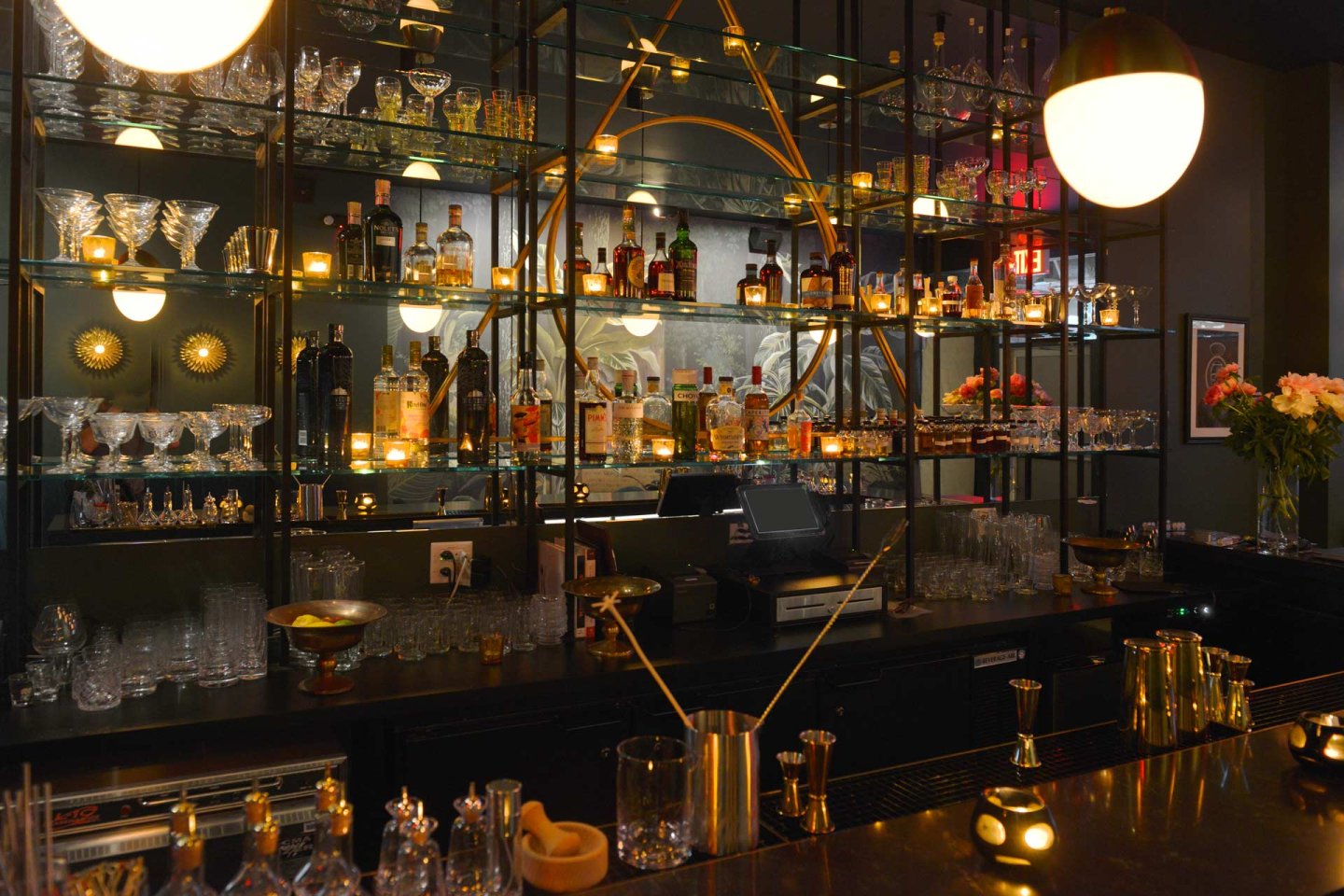 A dimly lit, elegant bar with mirrored shelves displaying various liquor bottles and glasses. Warm lighting creates a cozy ambiance, with decorative elements like flowers and candles enhancing the sophisticated atmosphere.