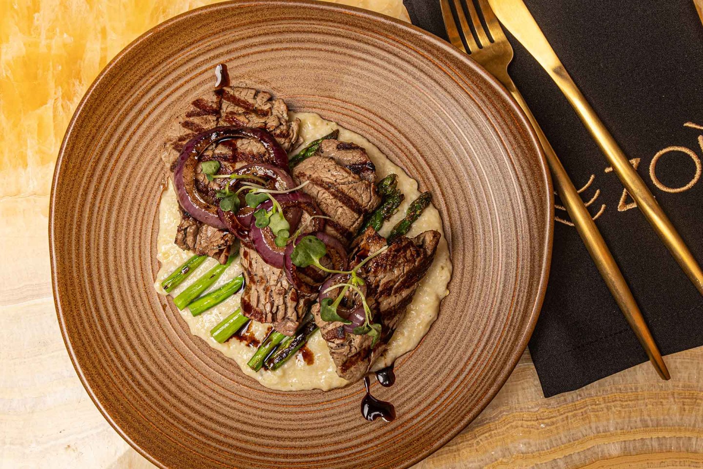 A plated dish with grilled steak slices, asparagus, red onion, and microgreens on a creamy sauce, served on a brown ceramic plate with gold-colored cutlery and a black napkin on a light wooden table.