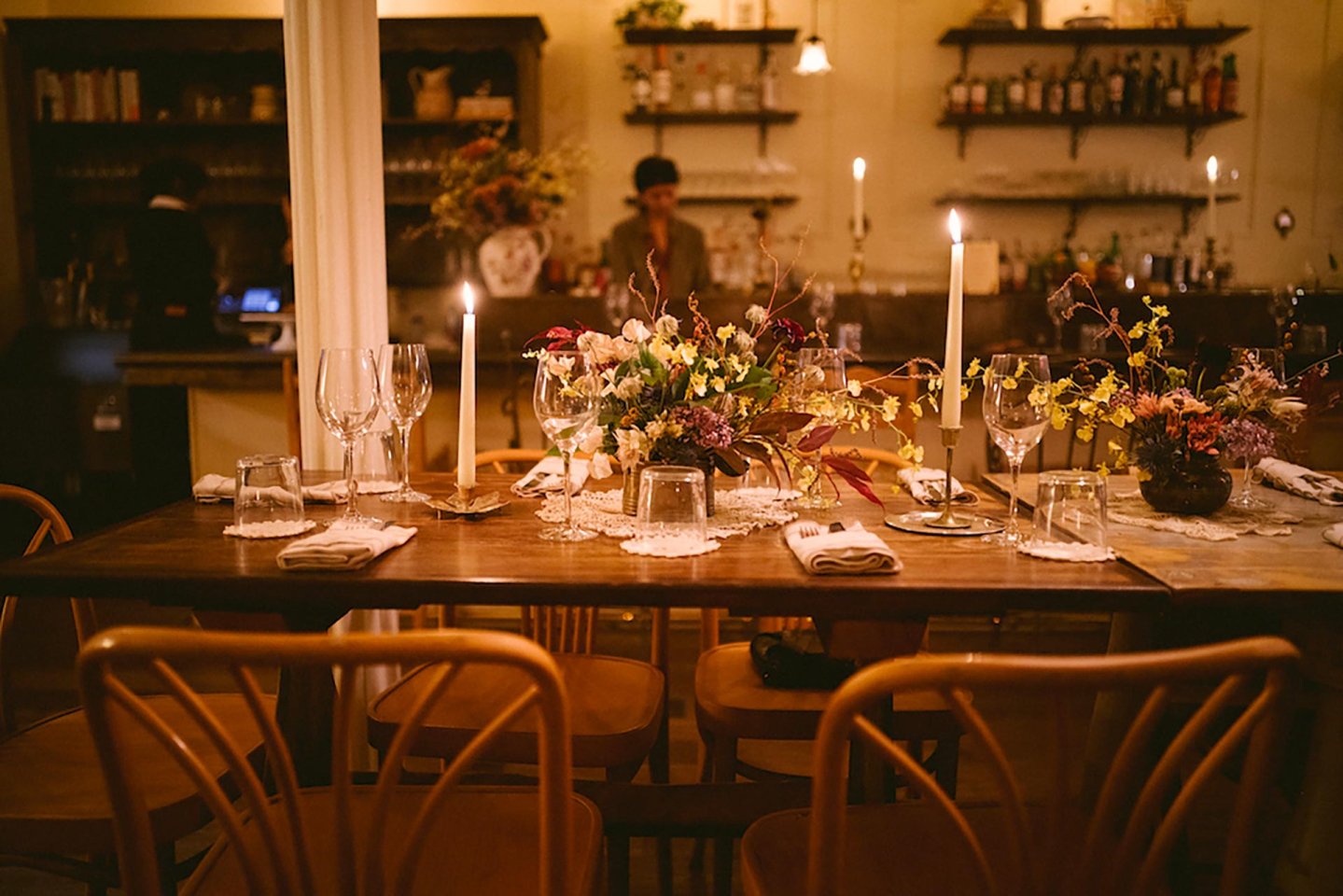 A warmly lit restaurant table set with candles, wine glasses, plates, and floral centerpieces. Wooden chairs surround the table, and shelves with bottles are visible in the background.
