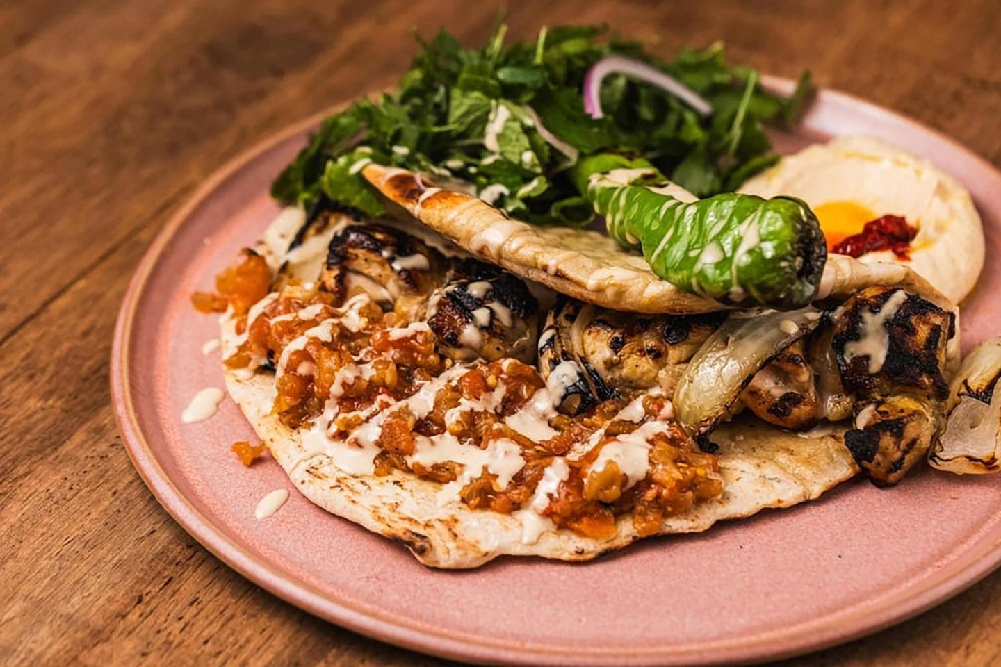 A plate with grilled chicken on flatbread topped with chopped tomatoes and white sauce, served with a grilled green pepper, fresh greens, onion slices, and a side of hummus, on a wooden table.
