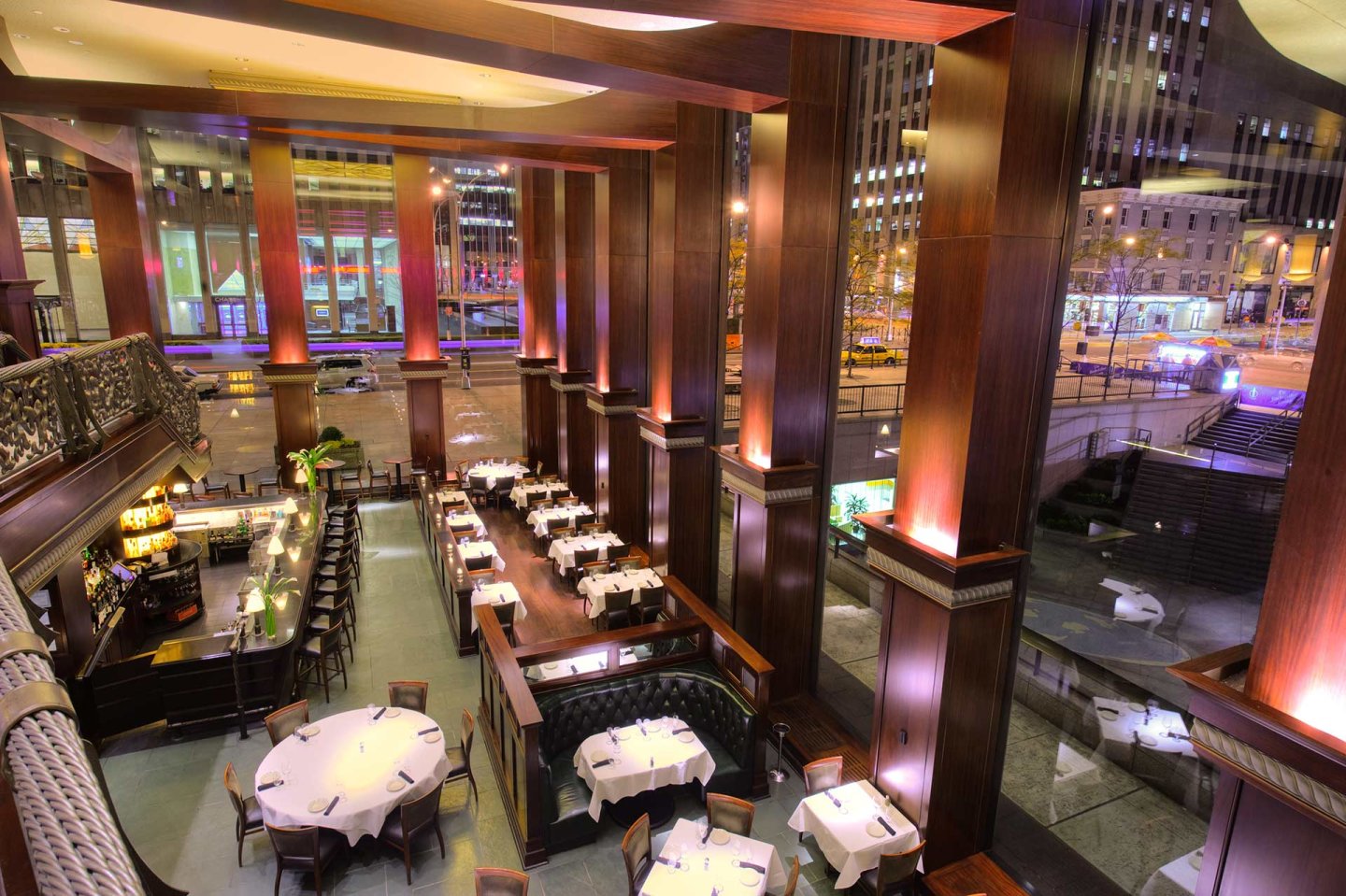 A spacious, upscale restaurant with high wooden columns, elegant lighting, white tablecloths, and large windows overlooking a brightly lit city street at night. Tables and booths are neatly arranged on two levels.