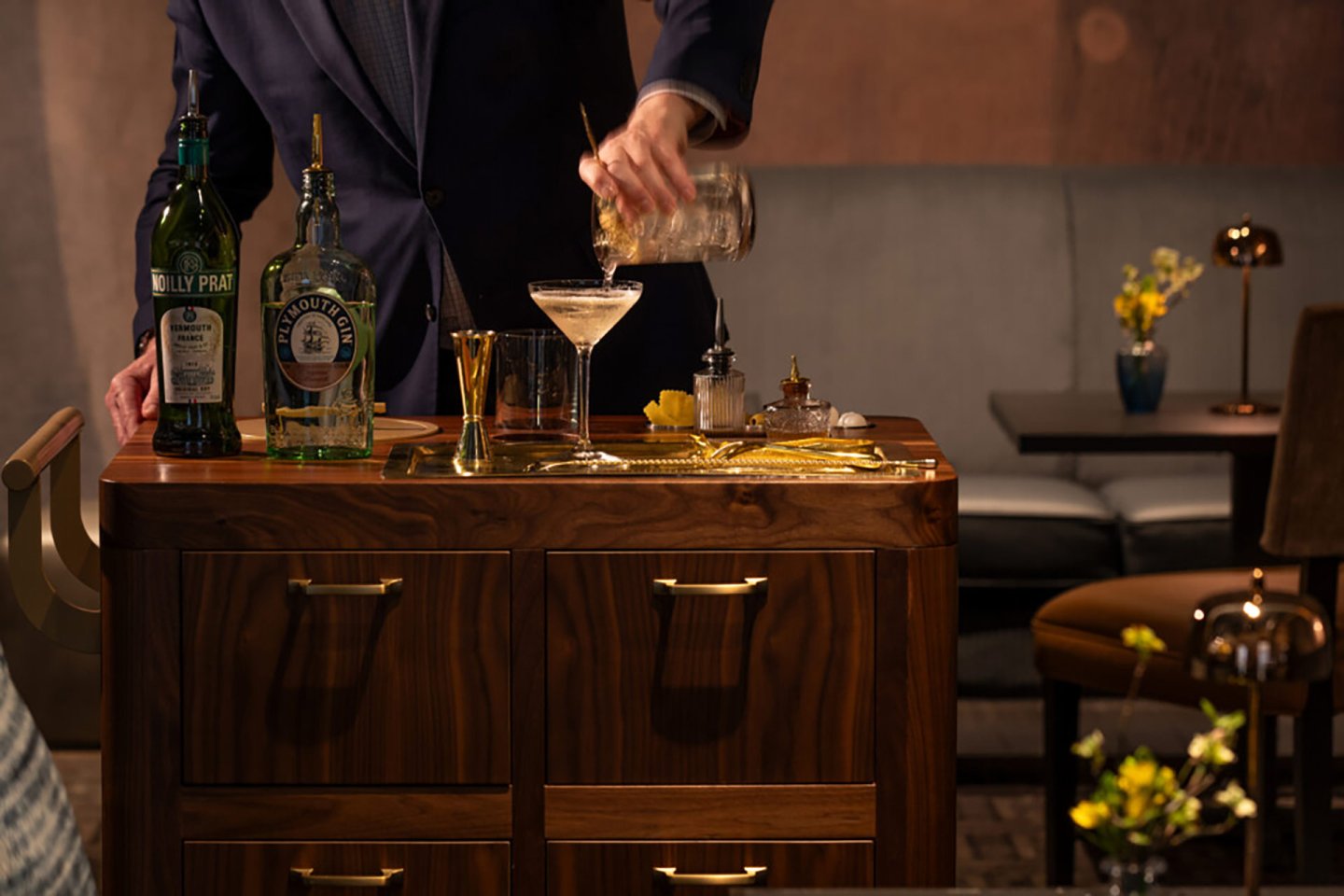 A bartender in a suit prepares a cocktail at a wooden bar cart with bottles, a martini glass, mixing tools, and flowers, in a warmly lit lounge setting.