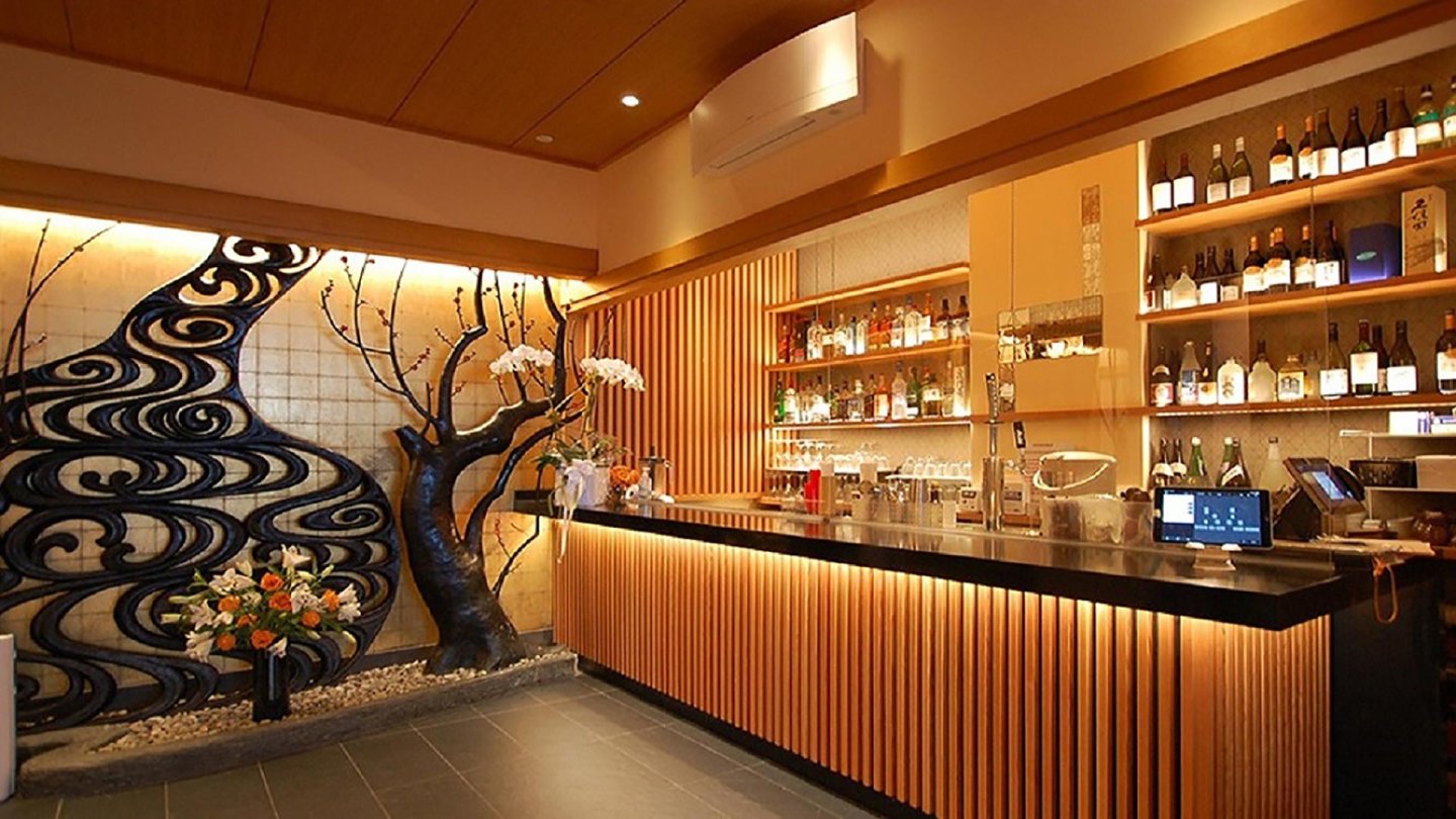 A modern Japanese bar with a black countertop, illuminated wooden slats, shelves stocked with bottles, and a decorative wall featuring a black tree sculpture and floral arrangements. Warm lighting creates a cozy atmosphere.