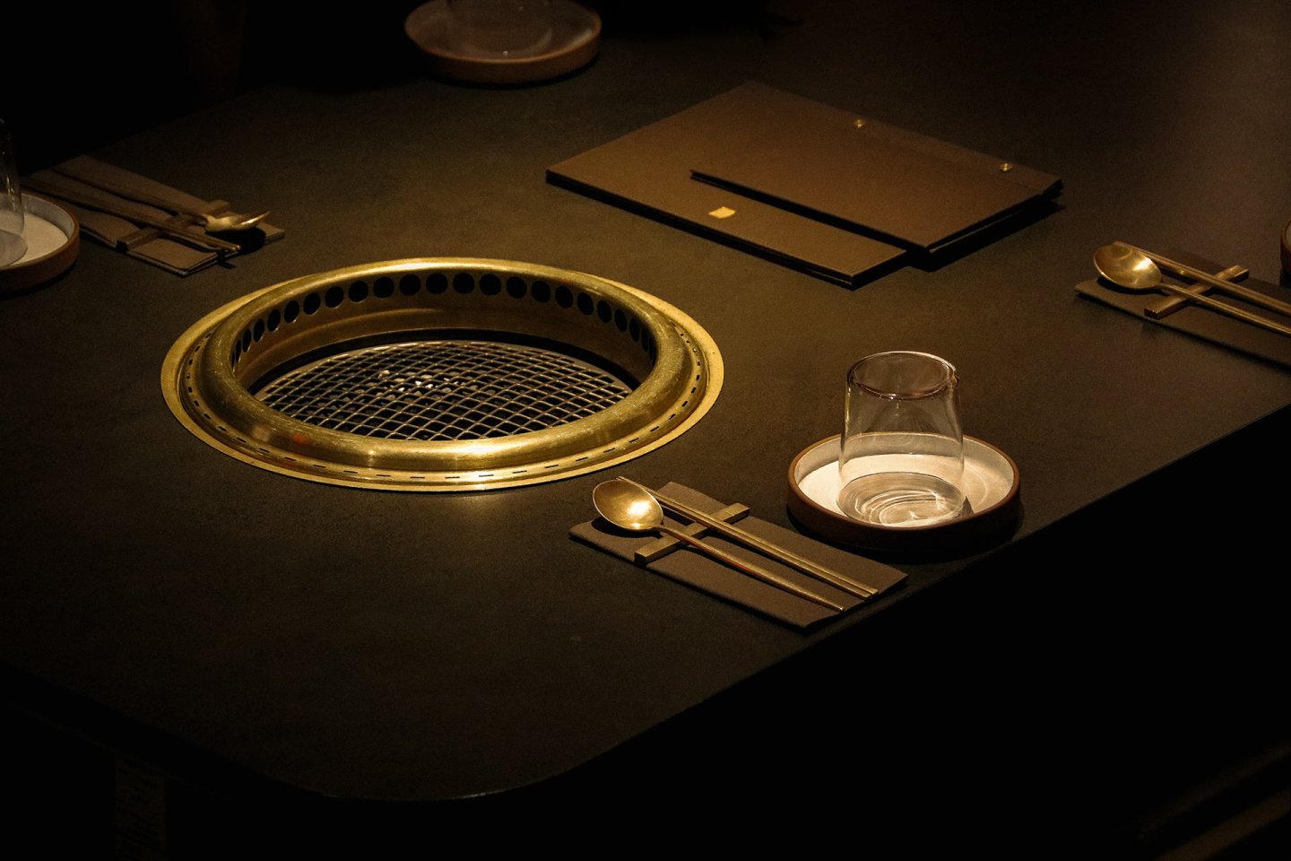 A dimly lit table set for dining features a built-in grill, utensils, plates, and glasses neatly arranged, creating an inviting atmosphere for a meal.