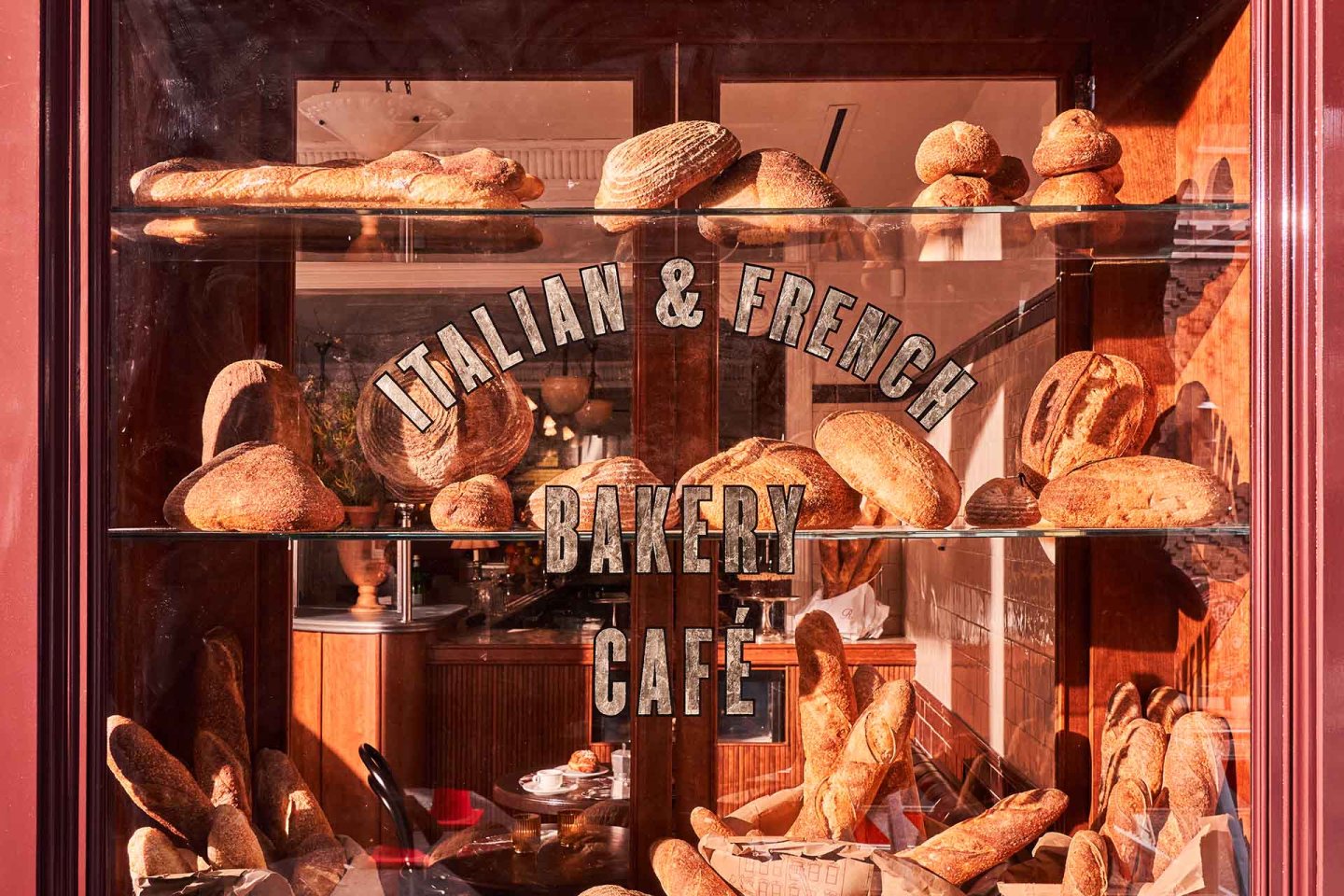 Bakery window displaying various artisanal breads on shelves. The glass reads "Italian & French Bakery Café." Inside, warm lighting and rustic wooden decor are visible.