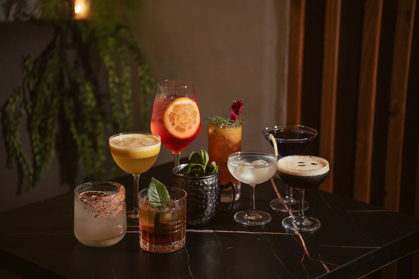 A variety of colorful cocktails in different glasses are arranged on a dark marble table, with garnishes like herbs and citrus slices. Green plants and wooden panels appear in the softly lit background.