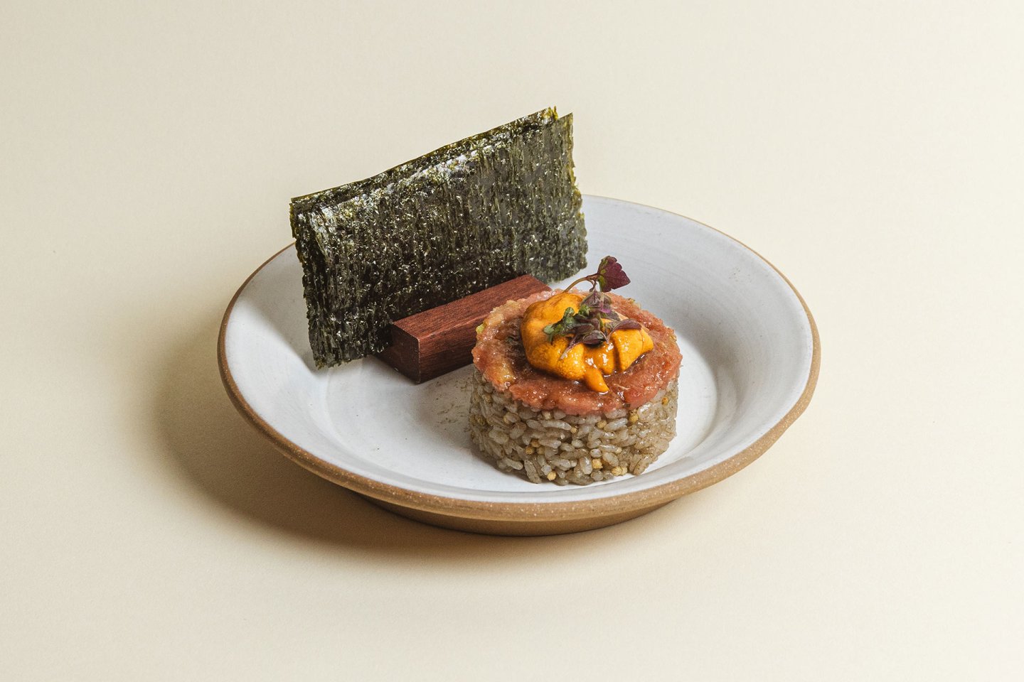 A plated dish featuring a round serving of rice topped with orange sea urchin and garnish, accompanied by a rectangular cracker and a sheet of nori, served on a white plate with a tan rim.