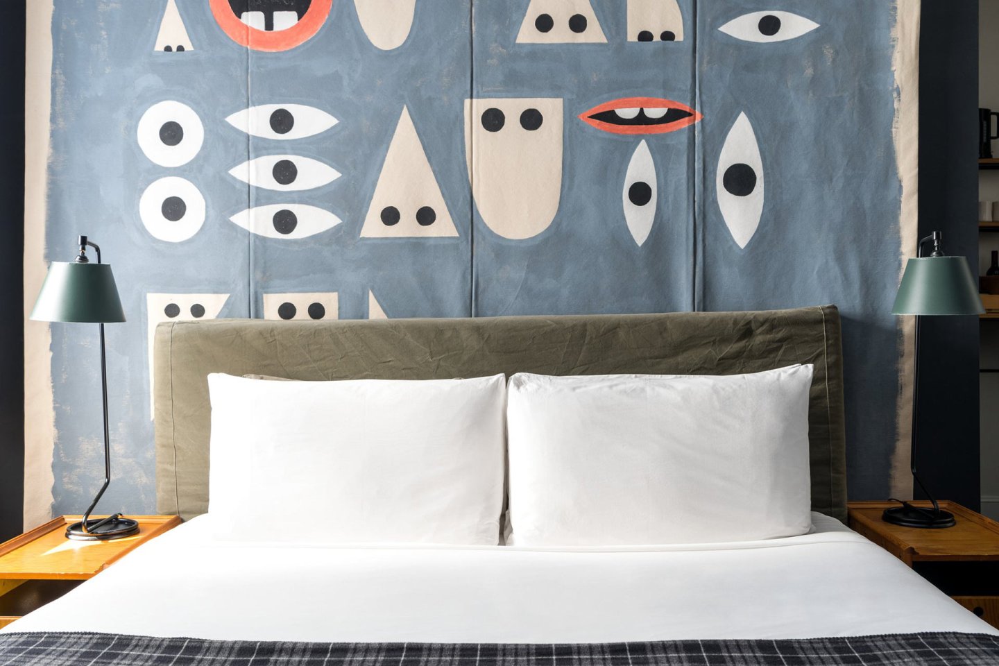 A neatly made bed with white pillows and sheets sits against a headboard. Behind it, a blue wall features abstract, geometric face designs. Two bedside tables each hold a green lamp.
