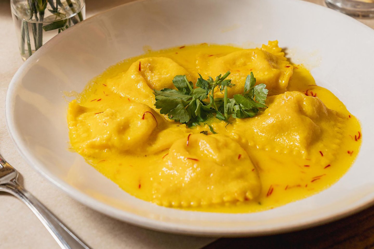 A white plate with five ravioli pieces in a creamy yellow saffron sauce, garnished with fresh parsley in the center. A fork and a glass are visible at the edges of the image.
