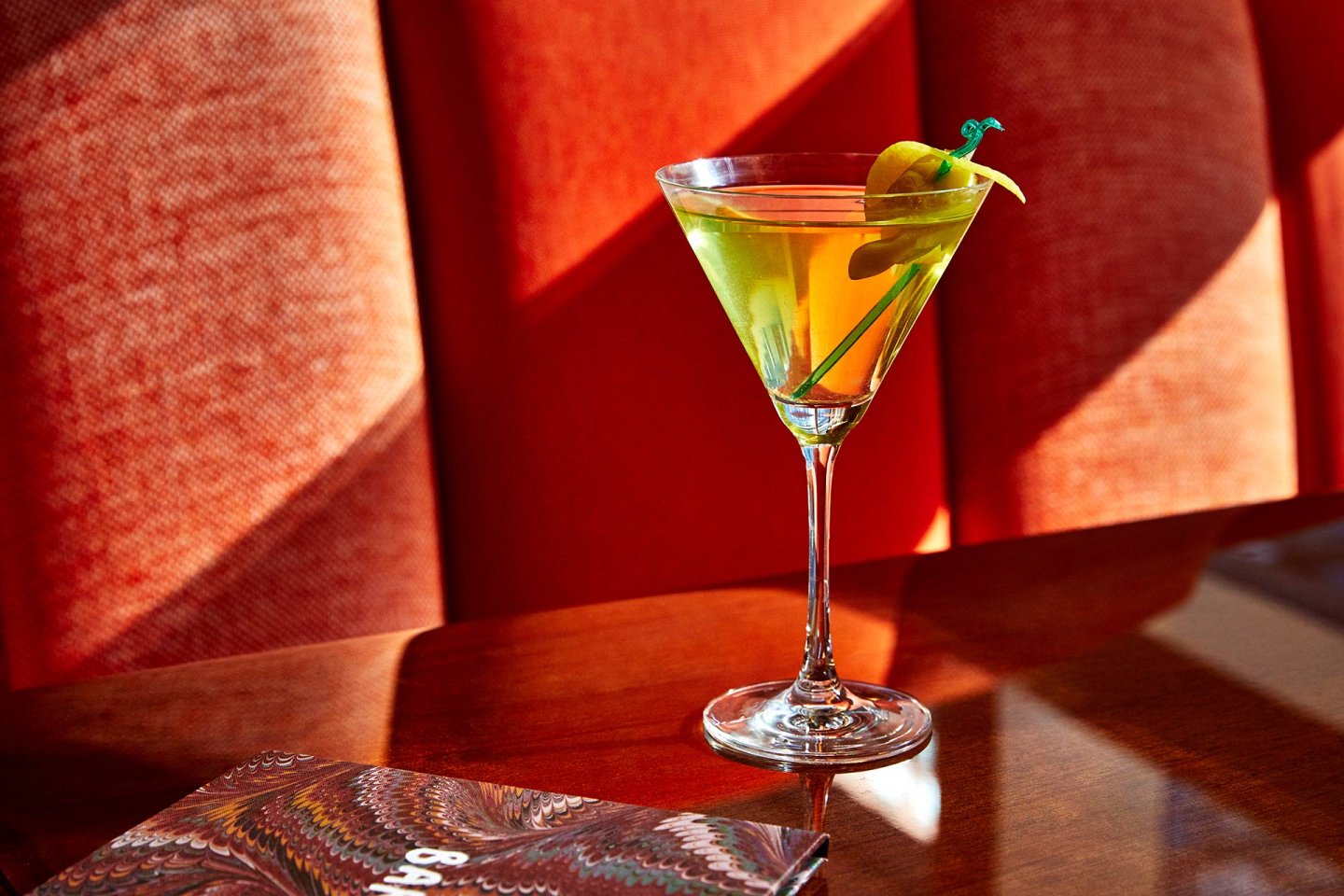 A martini glass with a green cocktail garnished with a twist of lemon and a green cocktail pick, sits on a shiny wooden table in front of an orange upholstered booth. A colorful menu is partially visible in the foreground.