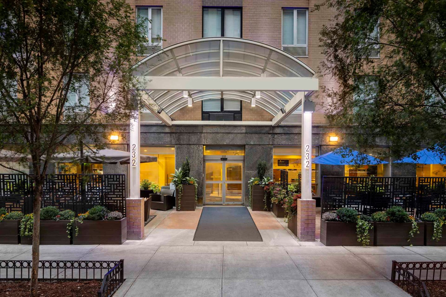 Exterior view of a building entrance with a transparent awning, large glass doors, and planters with greenery on either side. The entrance is flanked by address numbers "2332" on white beams.