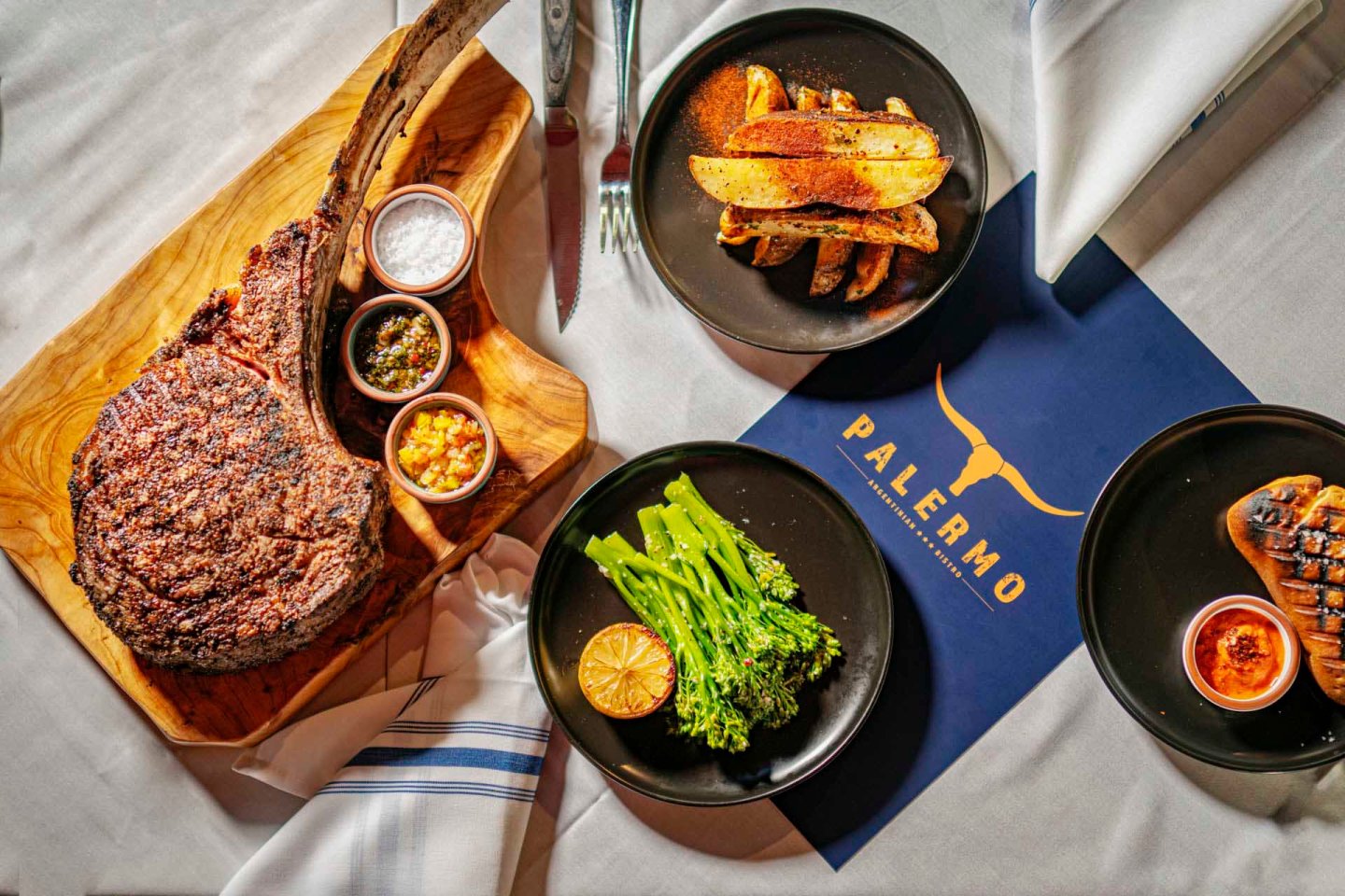 Tomahawk steak on a wooden board with small bowls of seasoning. Plates of potato wedges and broccolini arranged nearby on a table with a blue restaurant menu labeled "Palermo." A knife, fork, and napkin complete the setting.