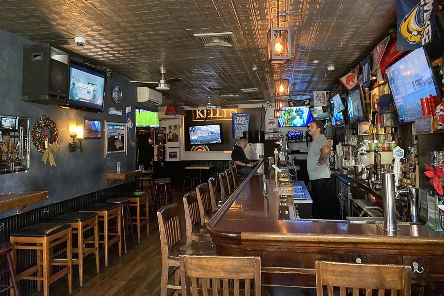 A cozy sports bar with wooden bar stools, multiple TV screens showing sports, two men behind the bar, and various sports memorabilia and flags decorating the walls and ceiling.