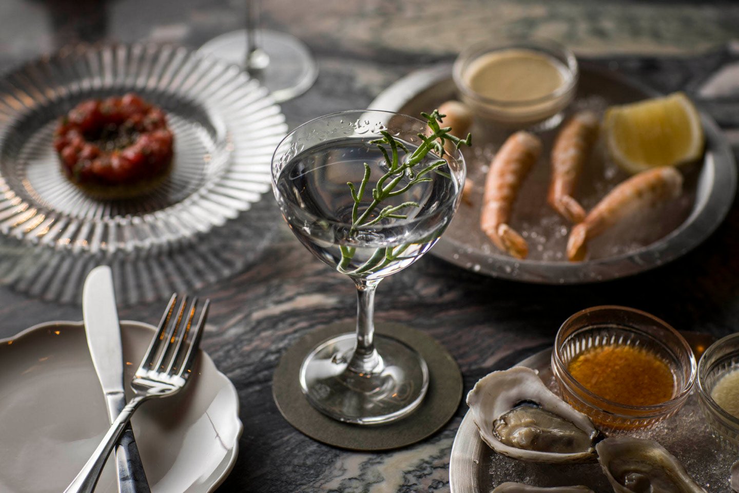 A dining table with a cocktail garnished with herbs, a plate of shrimp and lemon on ice, a dish of oysters with sauce, and a plate with a meat dish in the background. Cutlery is placed beside a white plate.