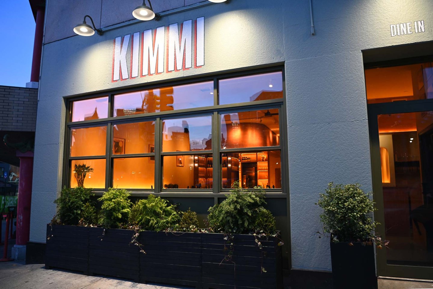 Restaurant exterior at dusk with large windows showing a warmly lit interior. "KIMMI" is displayed above the windows. Green plants in boxes line the front. "DINE IN" text is visible in the upper right corner.