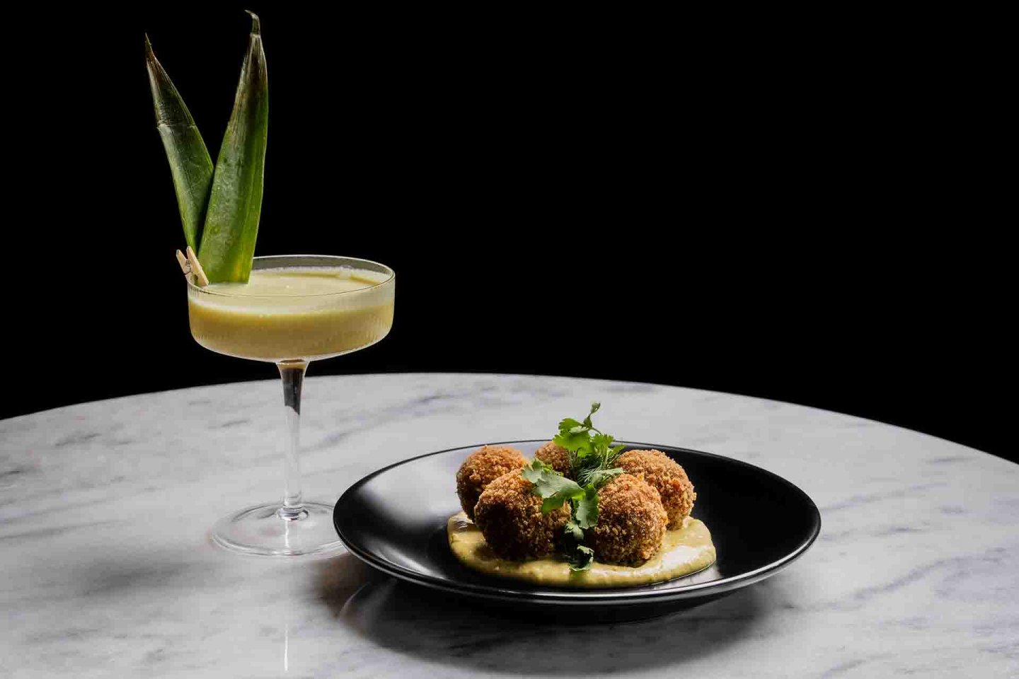 A cocktail garnished with pineapple leaves sits beside a black plate with three breaded croquettes on a creamy sauce, topped with fresh herbs, on a marble table against a black background.