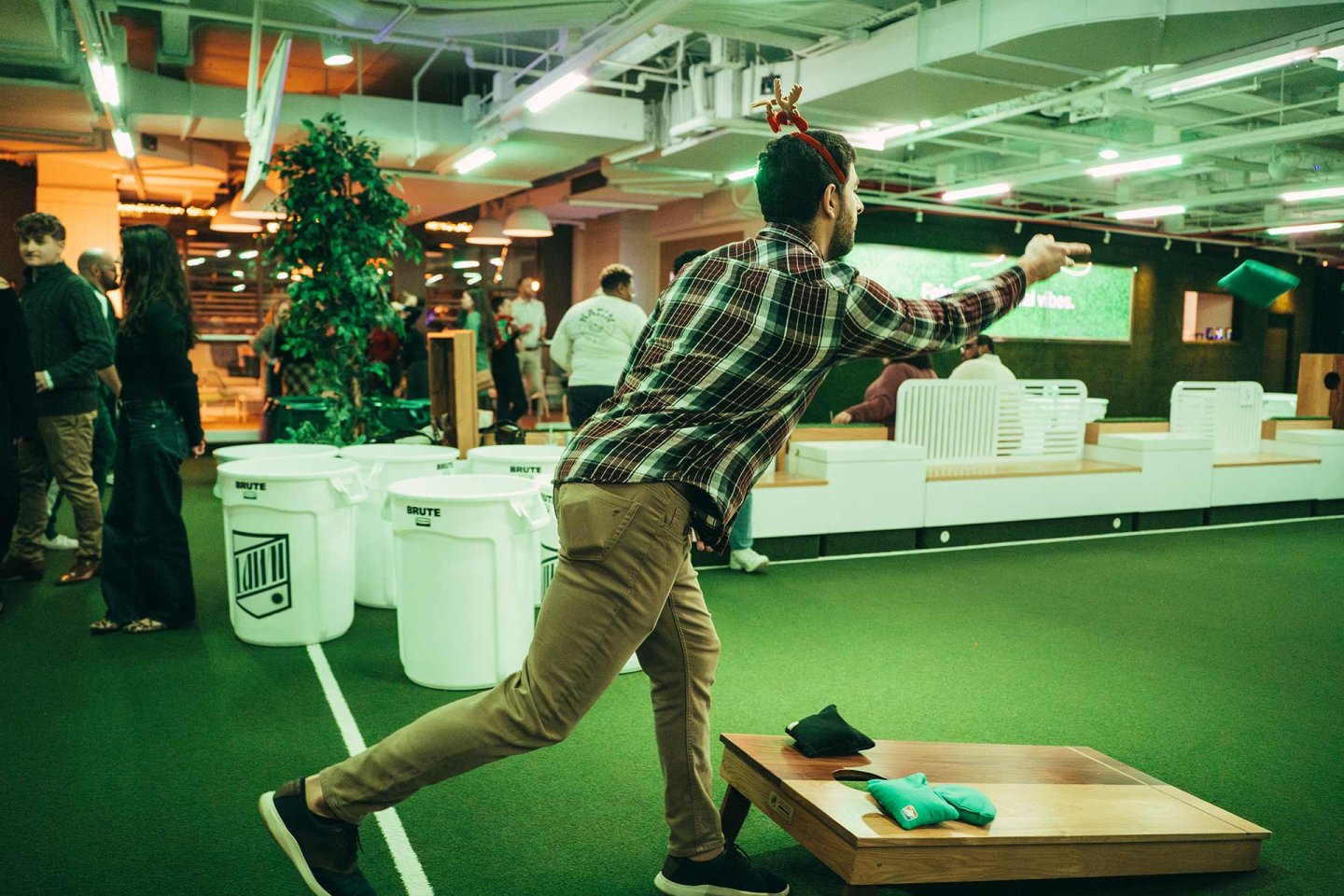 A man in a plaid shirt throws a bean bag while playing cornhole indoors. Other people are socializing around him on a green turf floor with benches and white decor.
