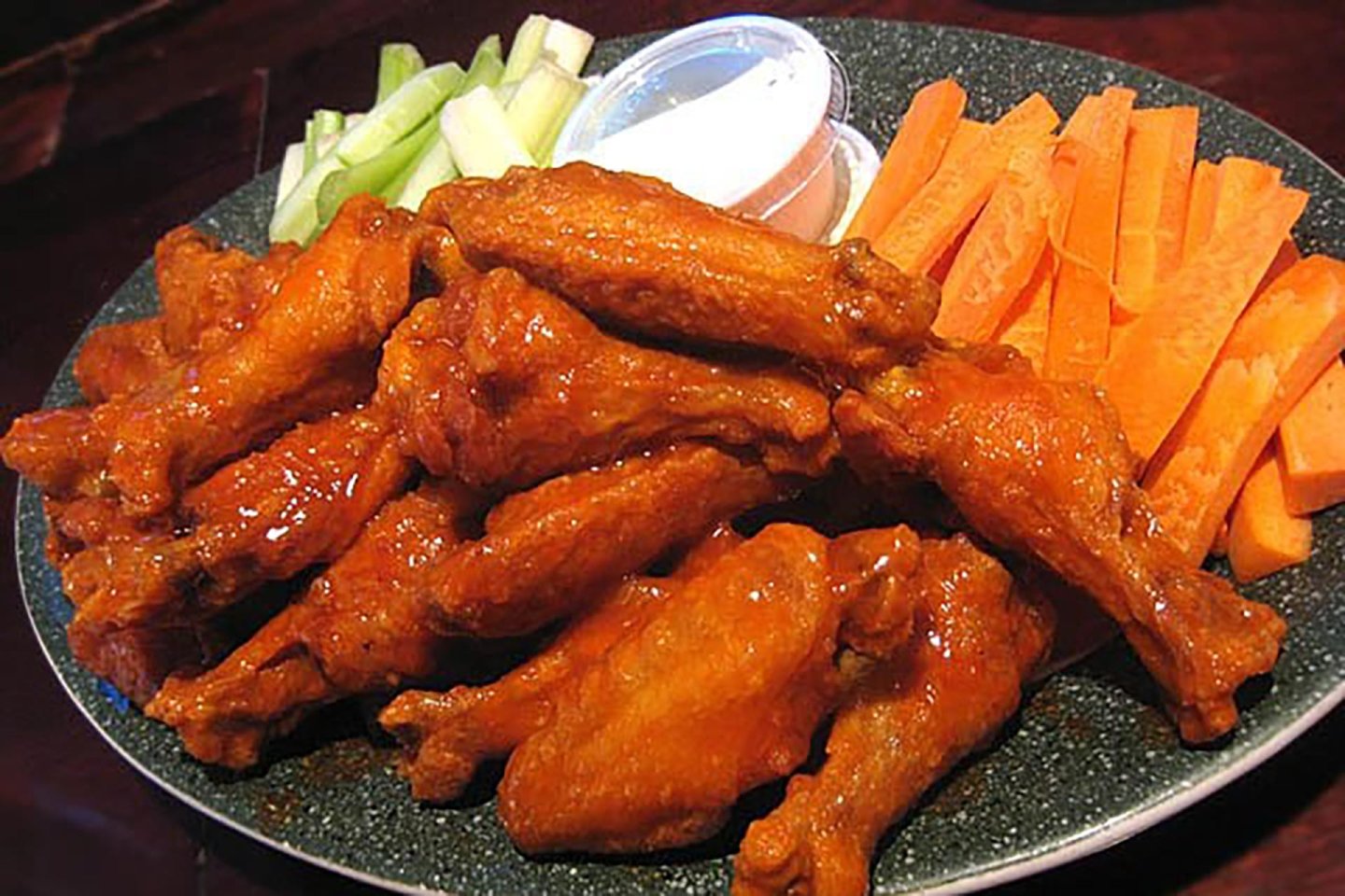A plate of saucy buffalo chicken wings is served with carrot sticks, celery sticks, and a cup of ranch or blue cheese dressing.