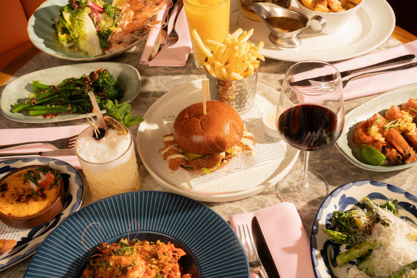 Restaurant table spread featuring a crispy chicken burger with fries, grilled prawns, salads, broccolini, pasta, cocktails, and a glass of red wine.
