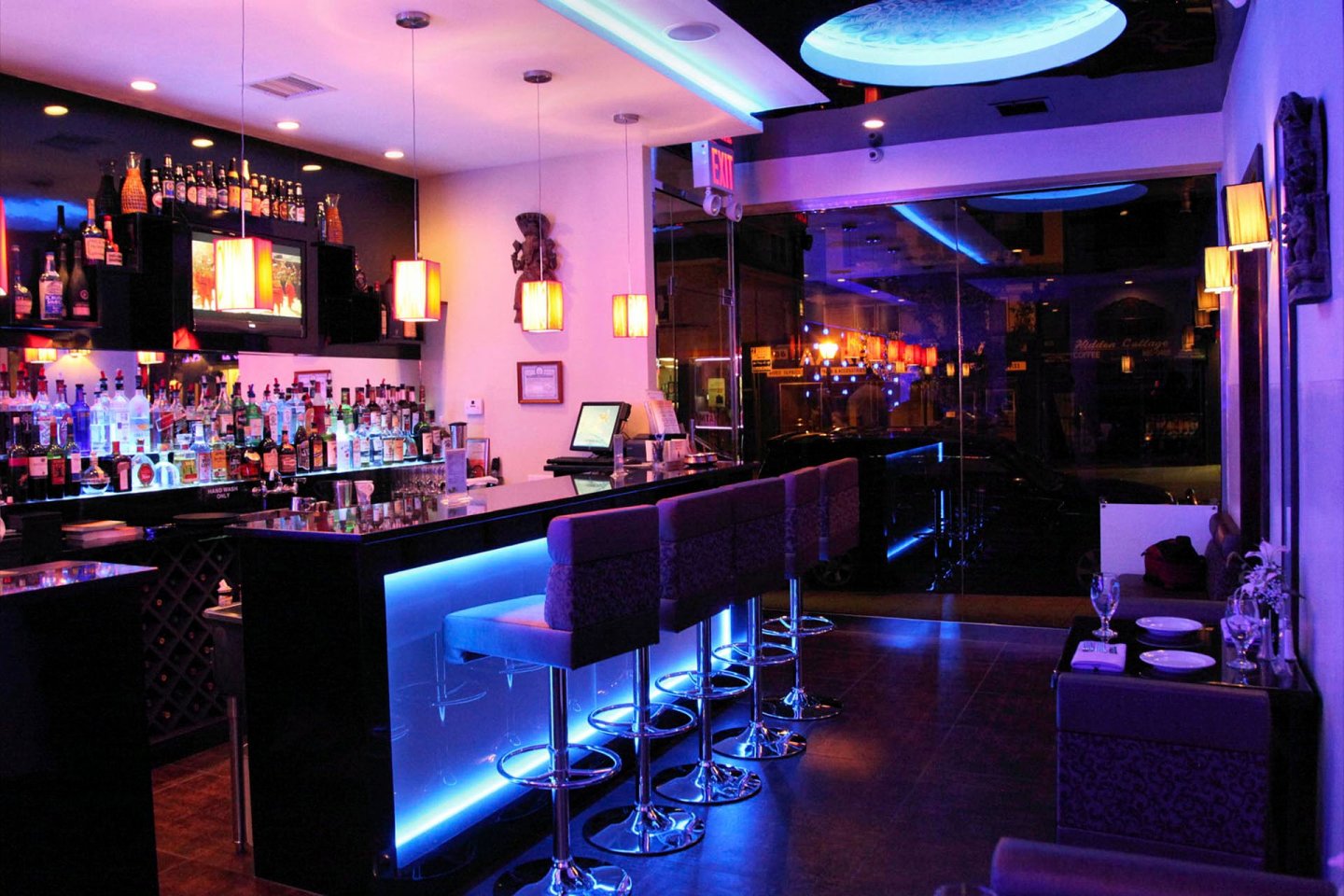 A modern bar with illuminated countertops, lined with high chairs. The bar is stocked with various bottles and glasses. Warm pendant lights hang above, creating an inviting atmosphere. The seating area is dimly lit with subtle blue accents.