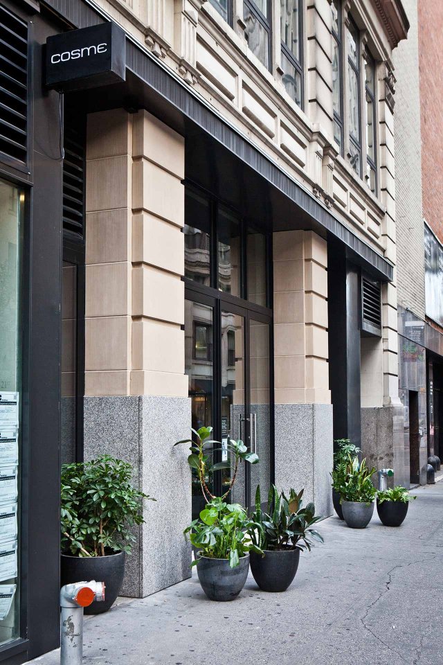 Street view of the entrance to a restaurant named Cosme, featuring large glass doors and several black planters with green plants arranged along the sidewalk. The building has beige stonework and tall windows.