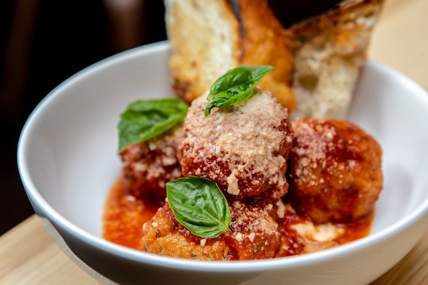 Three meatballs in tomato sauce topped with grated cheese and fresh basil leaves, served in a white bowl with slices of toasted bread in the background.