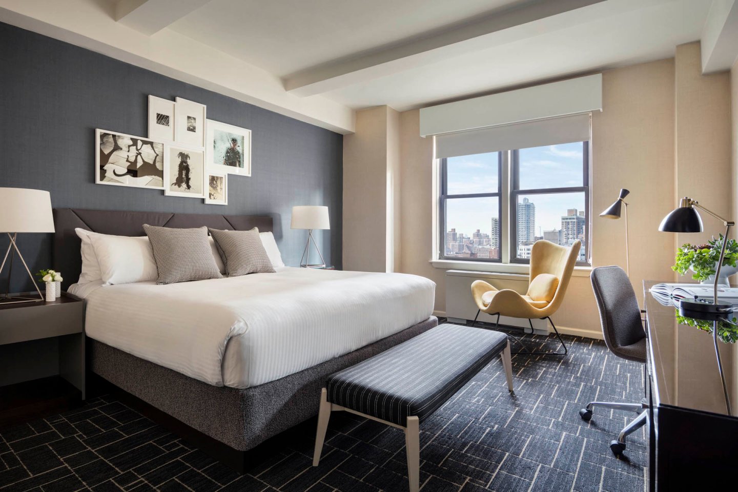 Modern hotel room with a large bed, gray and white bedding, a bench at the foot, a gallery wall of art, a yellow lounge chair by a window with city views, a desk with a lamp and chair, and two bedside lamps.