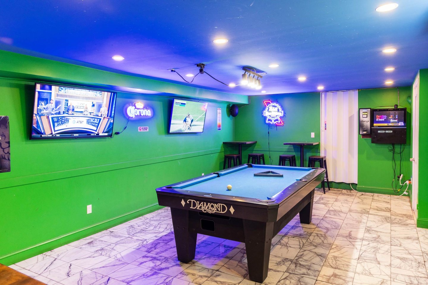 A brightly lit game room with green walls, marble tile floor, a pool table, two TVs showing sports, neon beer signs, bar tables with stools, and an electronic gaming machine in the corner.