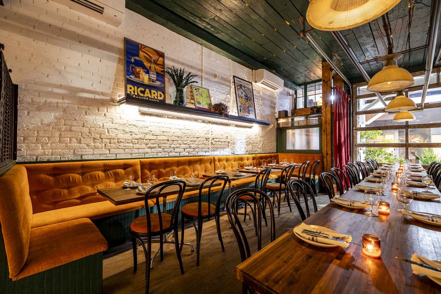 A cozy restaurant interior with long wooden tables, black chairs, cushioned orange bench seating, white brick walls, vintage posters, and warm pendant lighting. Place settings and lit candles decorate the tables.