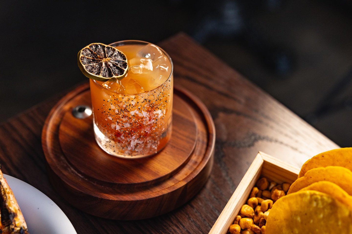 A cocktail with ice, chili flakes, and a dried citrus slice garnish sits on a round wooden coaster next to a tray of snacks, including yellow chips and roasted corn.