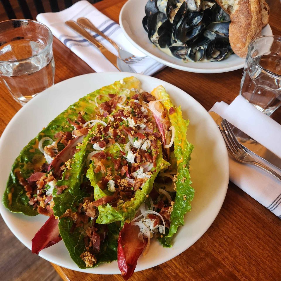 A plate of fresh romaine lettuce salad topped with bacon, onions, and crumbled cheese. In the background, a bowl of mussels with bread, glasses of water, and napkins with utensils on a wooden table.