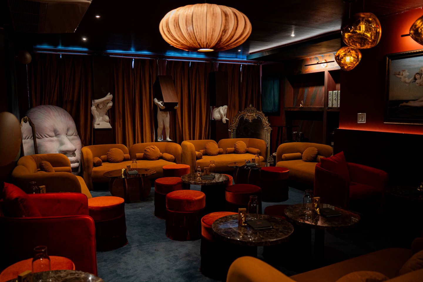 A dimly lit lounge with orange and red seating, round marble tables, modern art sculptures, a large decorative mirror, and warm hanging lamps, creating a cozy, artistic atmosphere.