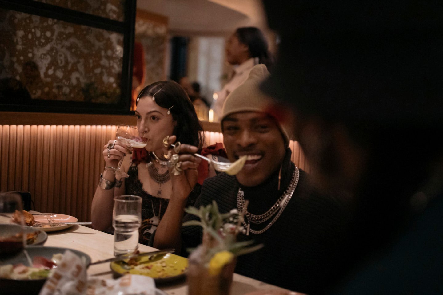 A group of people seated at a dimly lit restaurant table, enjoying their meal. In the foreground, a person wearing a cap and jewelry is eating, while a person with dark hair and hair clips sips from a glass. Plates and drinks are on the table.