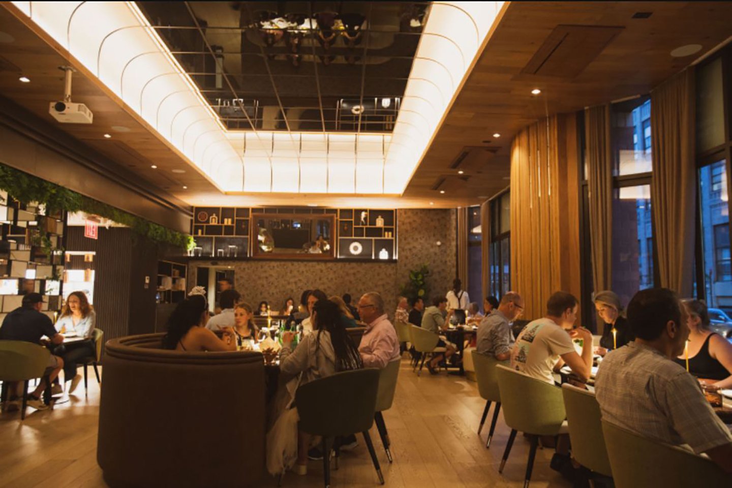 A warmly lit, modern restaurant with people dining at tables, wooden floors, tall windows, and a stylish ceiling light fixture create a cozy, lively atmosphere.