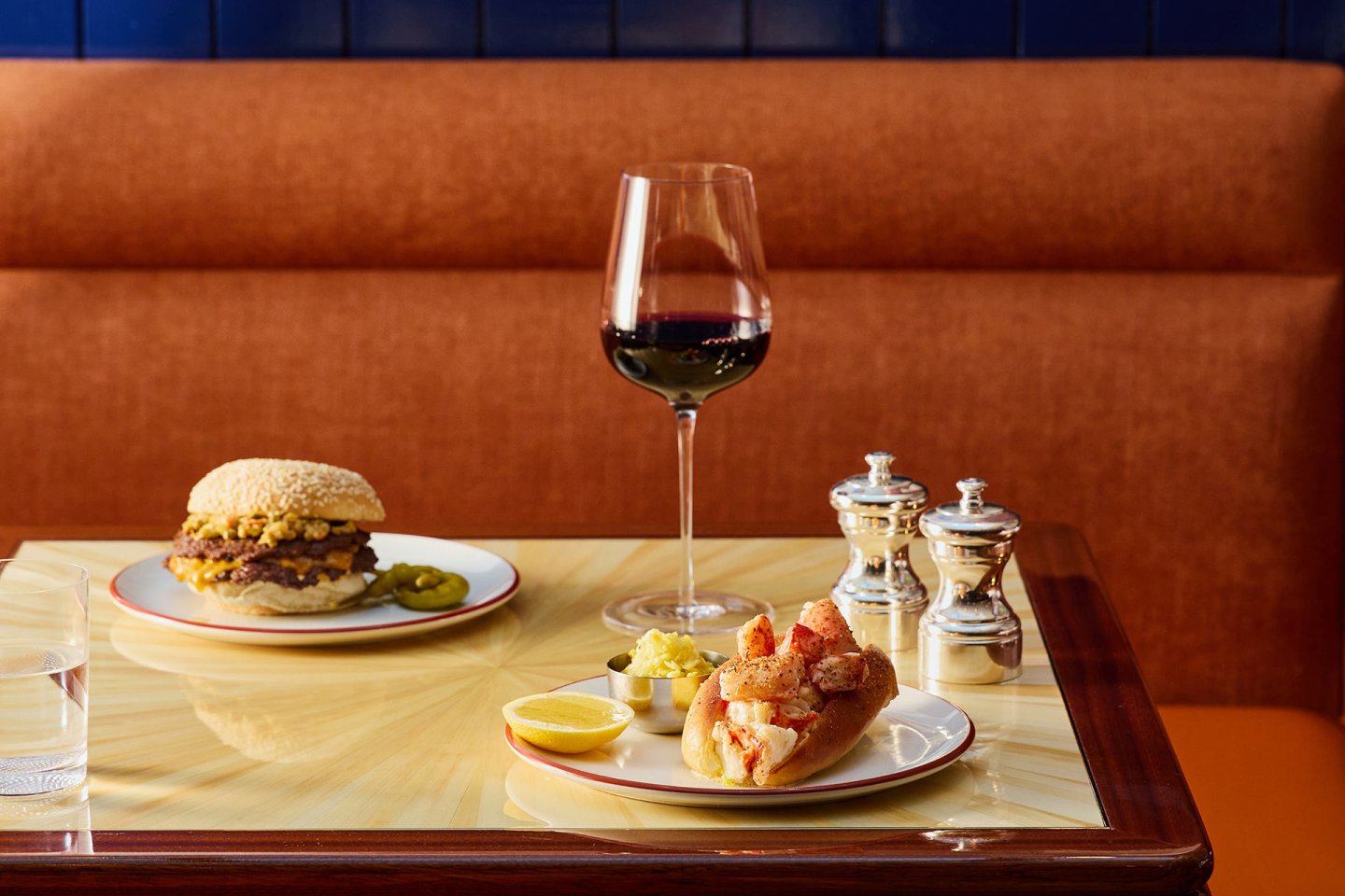 A table with a burger, a lobster roll with lemon, a glass of red wine, salt and pepper shakers, and a small dish of pickles on a yellow surface with a brown upholstered bench in the background.