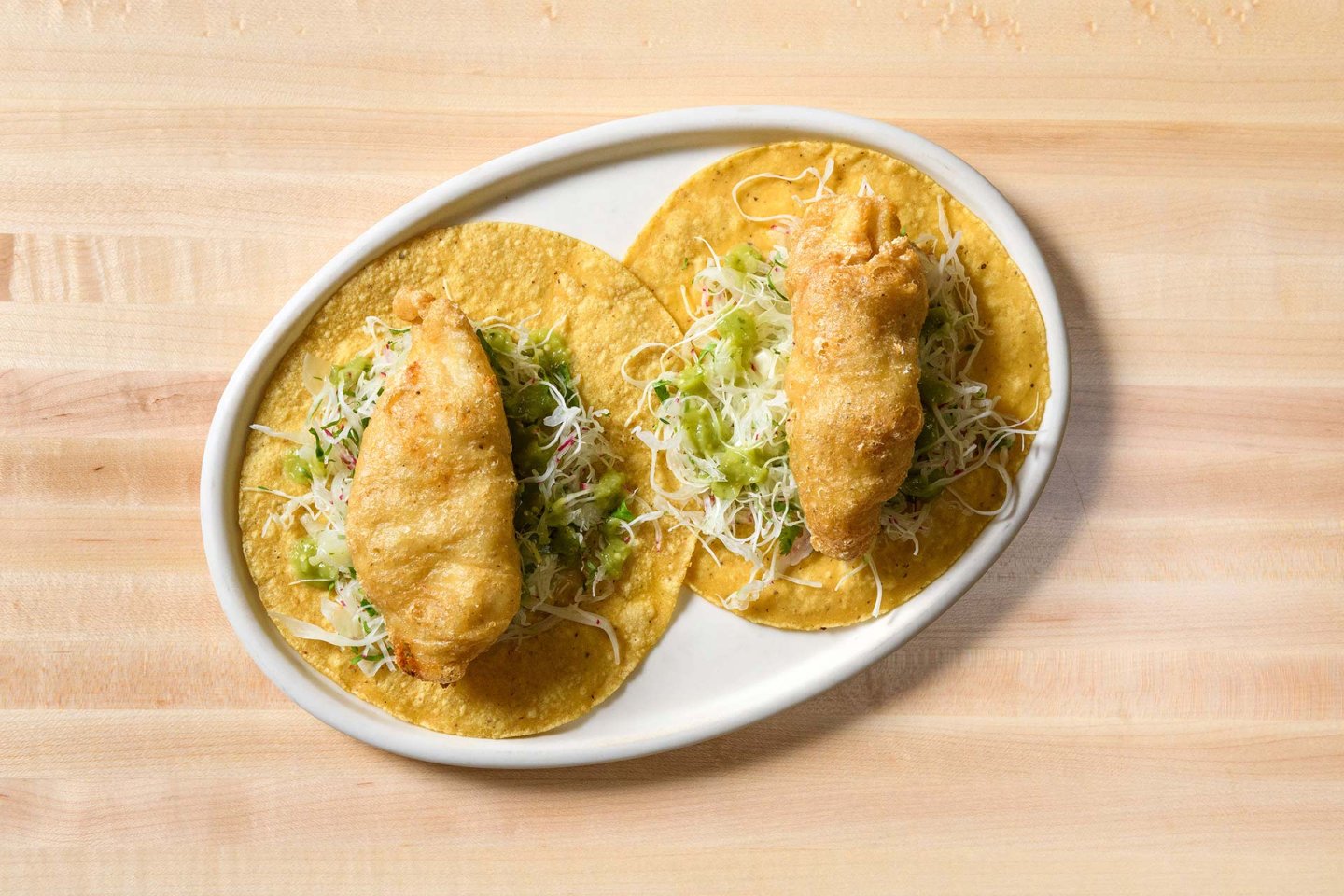 Two crispy battered fish tacos topped with shredded cabbage and cheese, served on corn tortillas on a white oval plate, placed on a light wooden table.