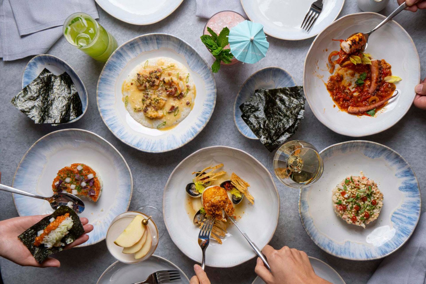 A table with various plated dishes, including seafood and pasta, accompanied by seaweed, sliced apples, and drinks. Hands hold forks, reaching for food. The setting suggests a shared dining experience.